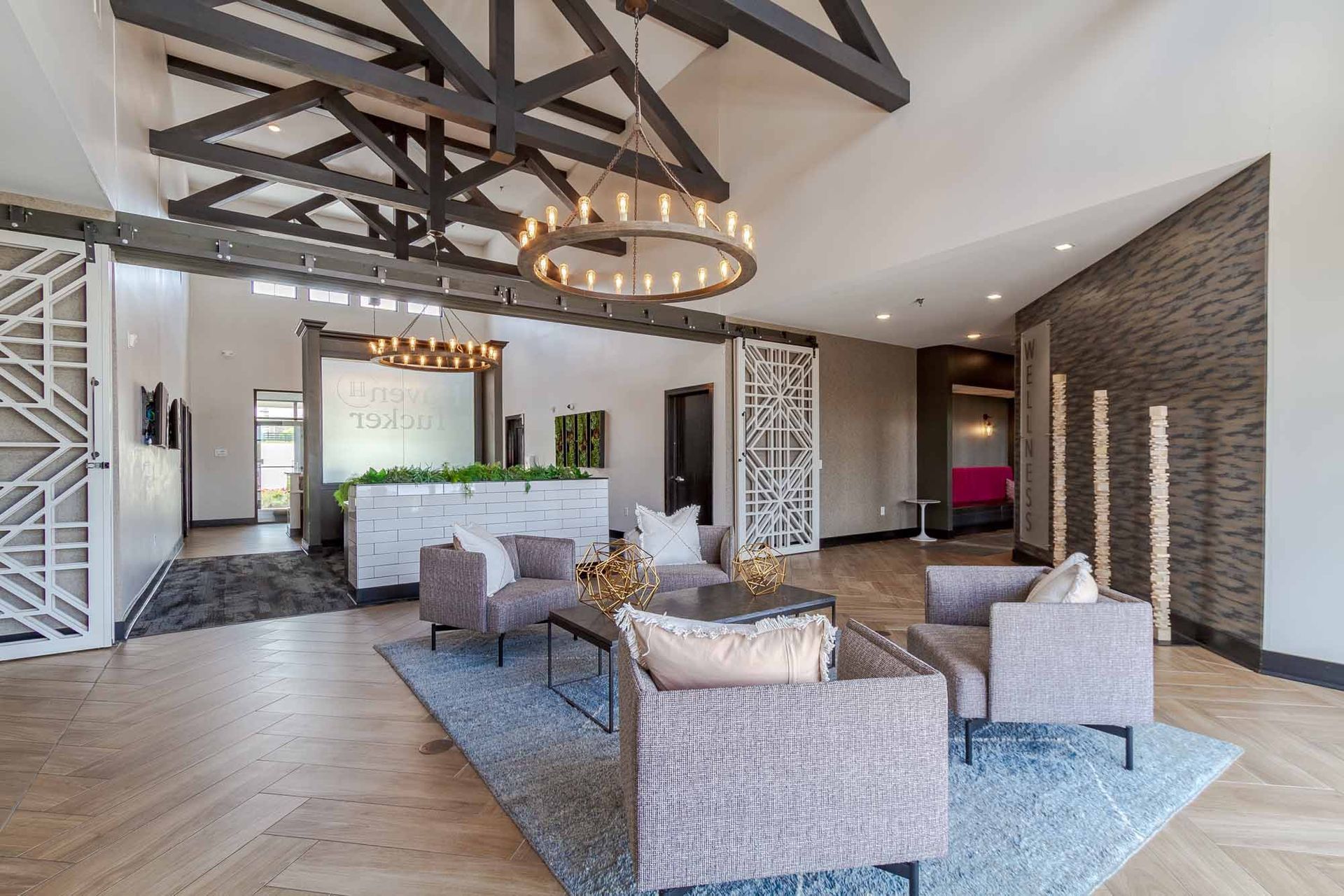 Lobby with seating, exposed beams, and a large chandelier. Light wood floors, blue rug, and modern decor.