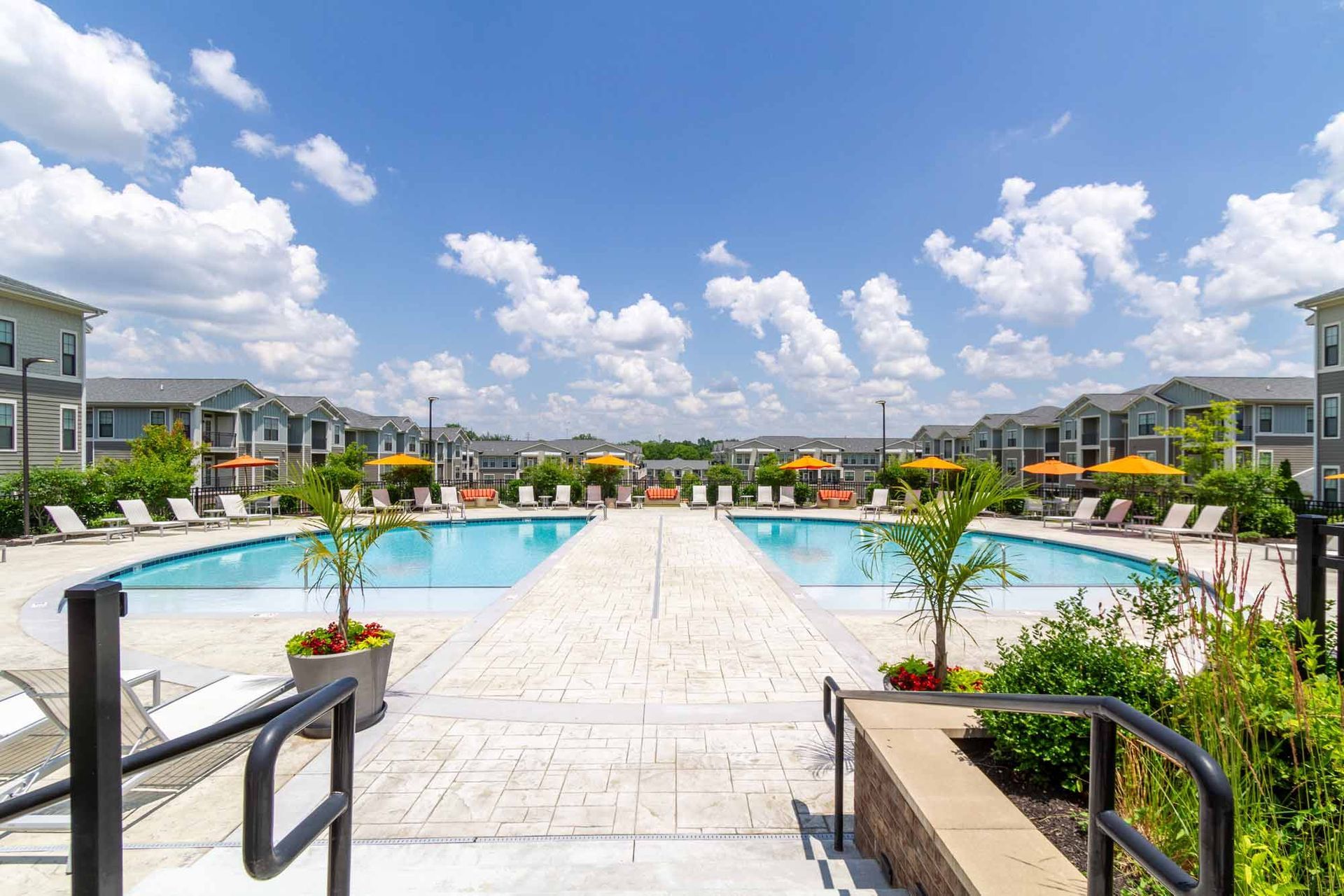 Two symmetrical pools with lounge chairs, surrounded by apartment buildings under a blue sky with clouds.