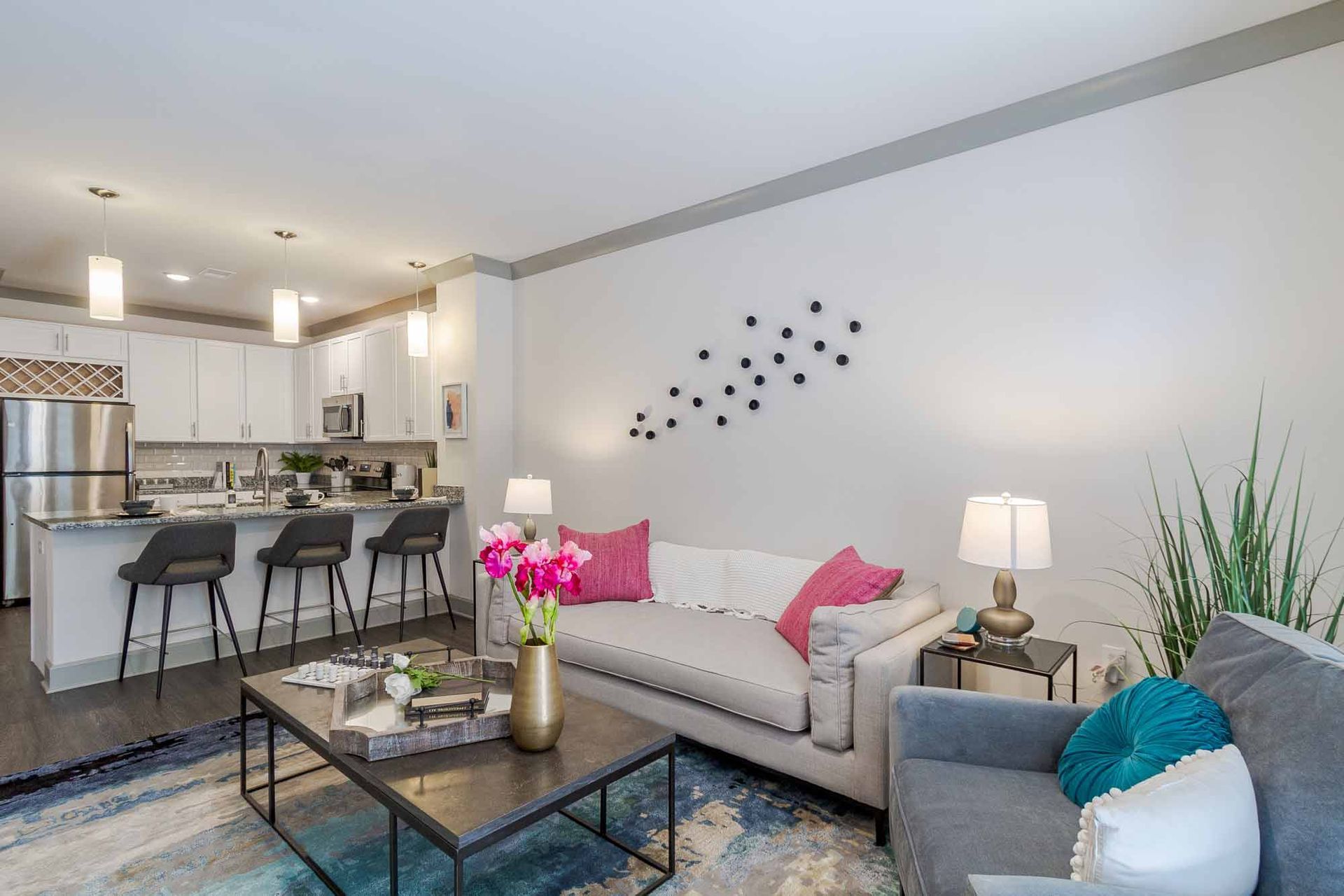 Open-concept living space with kitchen and seating area. White walls, grey trim, neutral sofa with pink pillows, and a patterned rug.