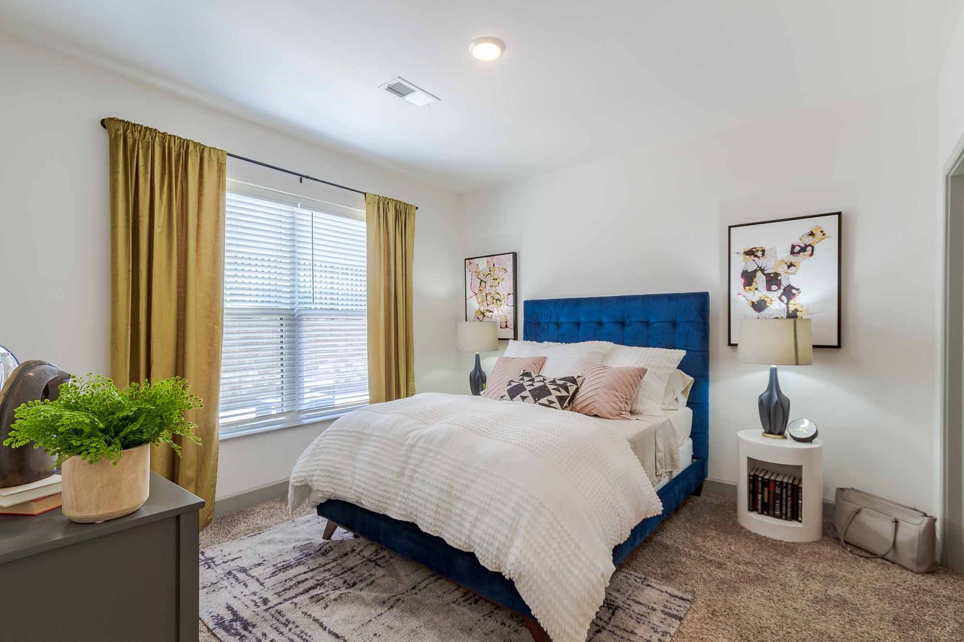 Bedroom with blue velvet bed, white bedding, gold curtains, and a plant on a gray dresser.