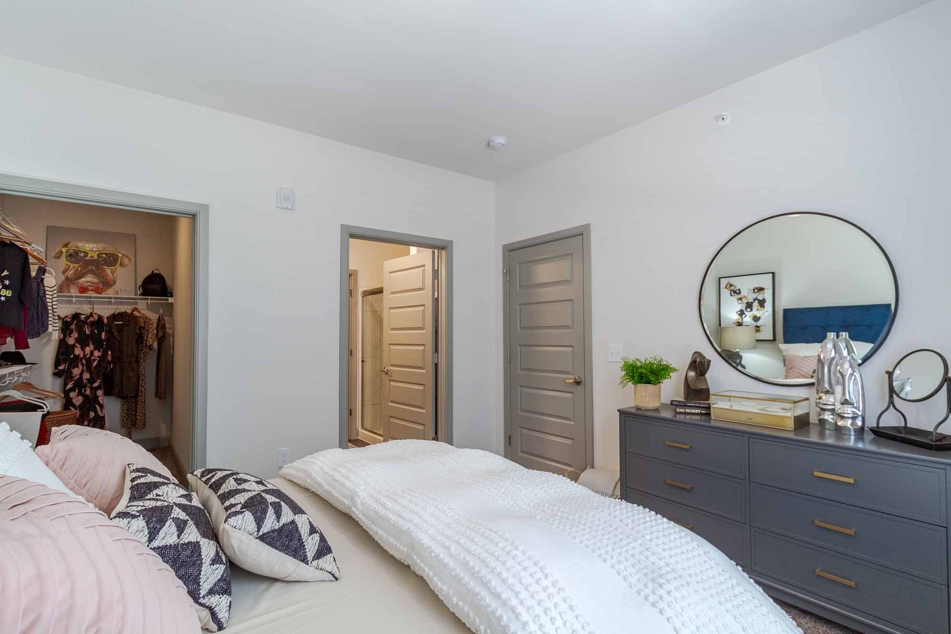 Bedroom with bed, dresser, closet, and doors; light gray walls, white bedding, and a round mirror.