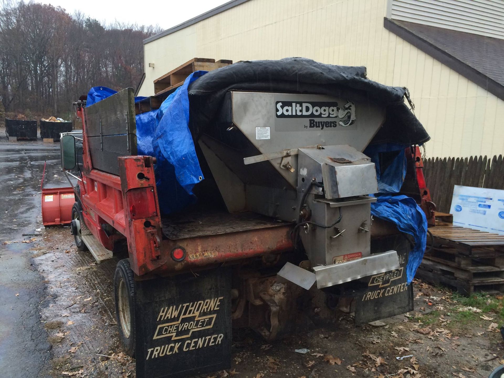 Red truck with a salt spreader covered by a blue tarp. Truck is parked outdoors.