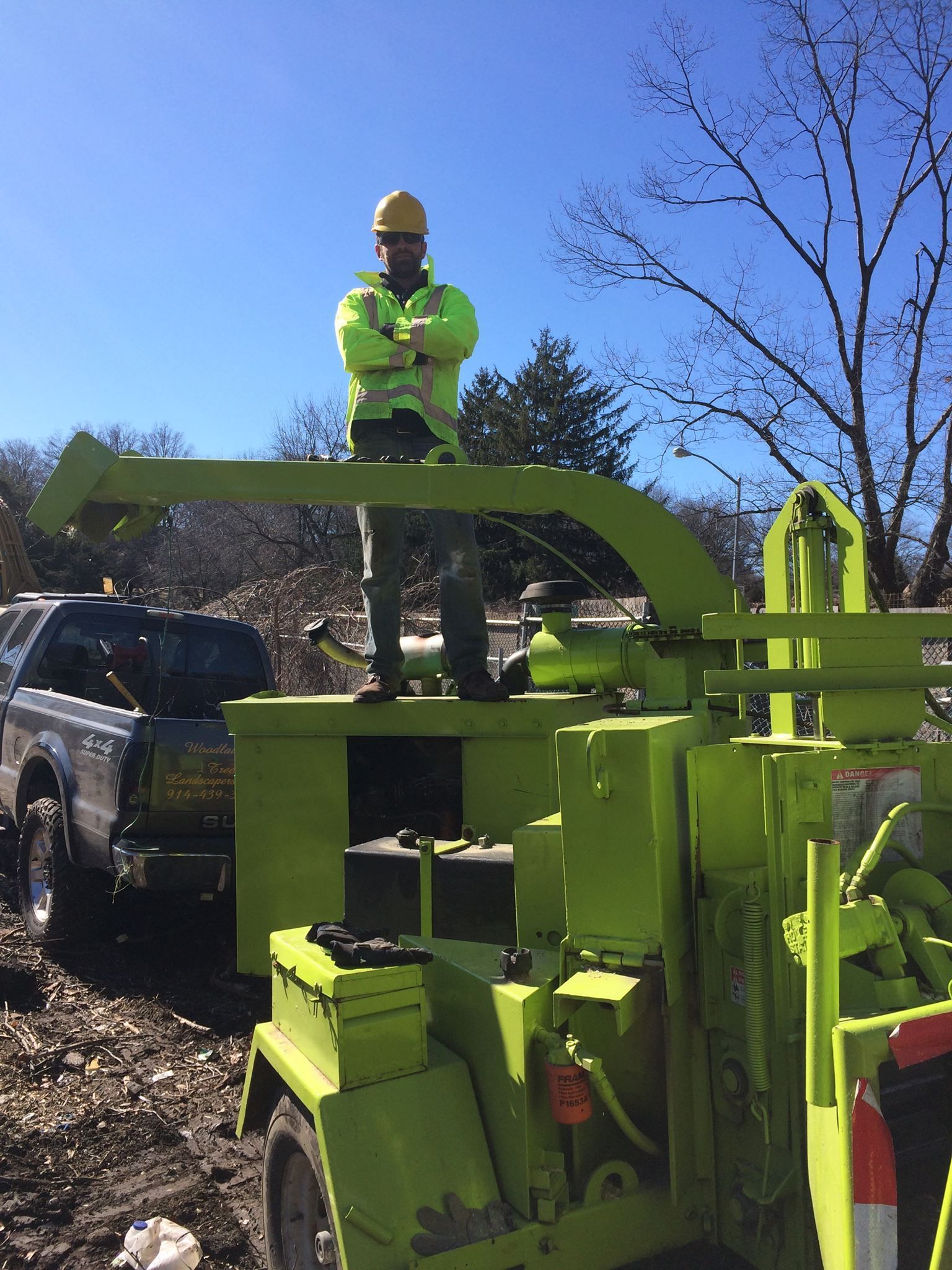 Man in safety vest and hard hat stands atop a green wood chipper, arms crossed, near a truck.