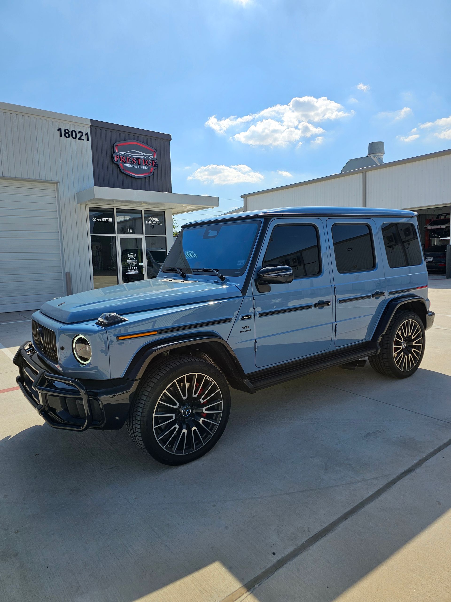 Blue Mercedes-Benz G-Class SUV parked on concrete in front of a building with a business sign.