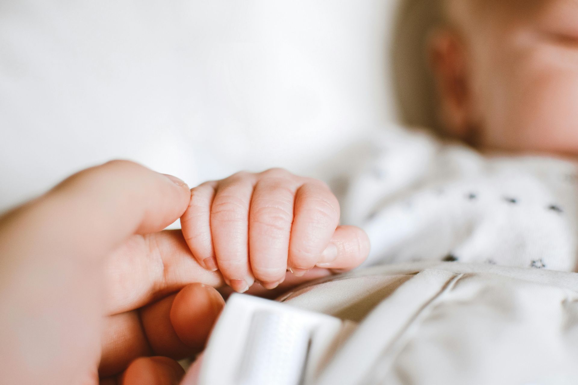 Close-up of a baby’s hand holding an adult finger on a white blanket