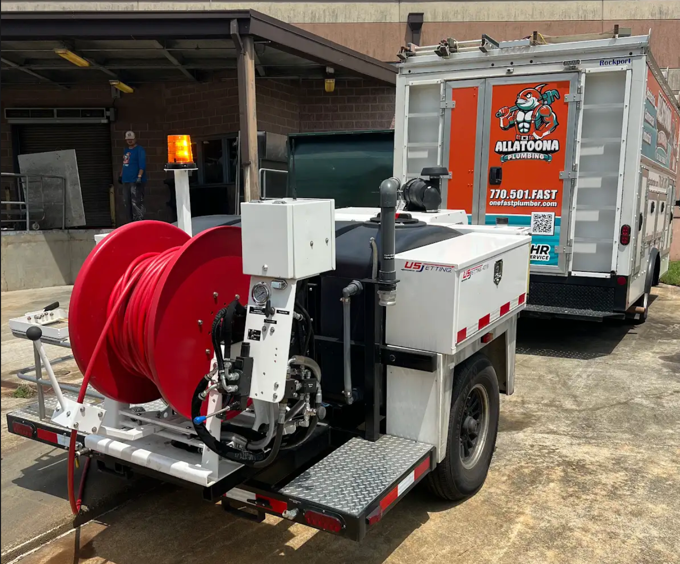 A white trailer with red hose reel, next to a truck with orange logos, outside a building.