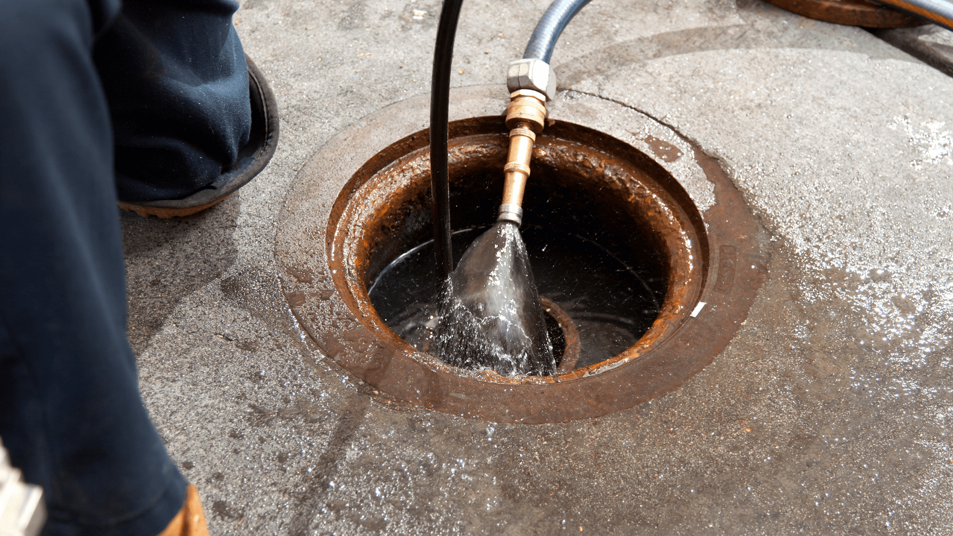 Person using a hose to flush a manhole with water on a concrete surface.