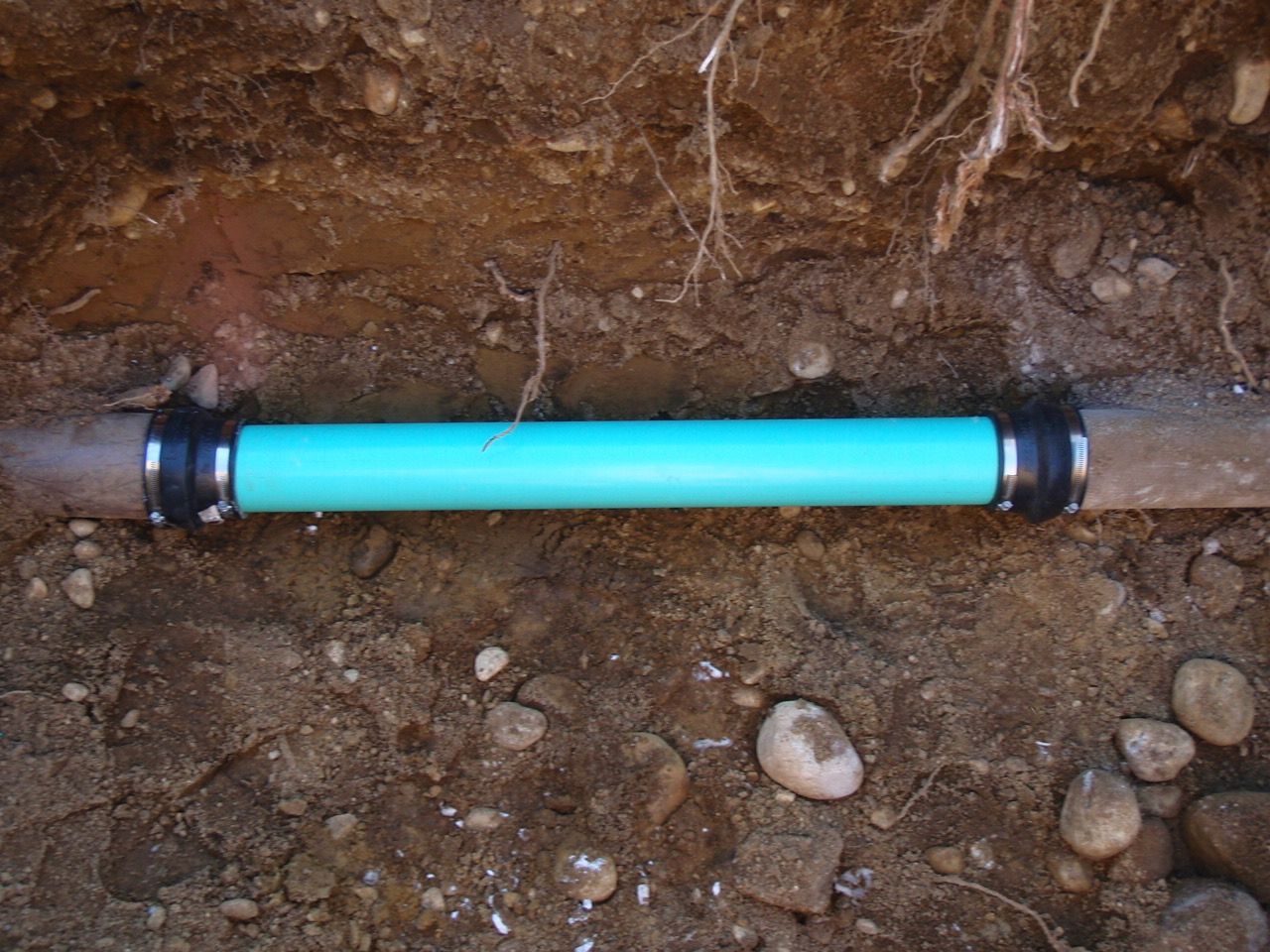 A blue PVC pipe section connects two brown pipes in a dirt trench, fixed with black couplings.
