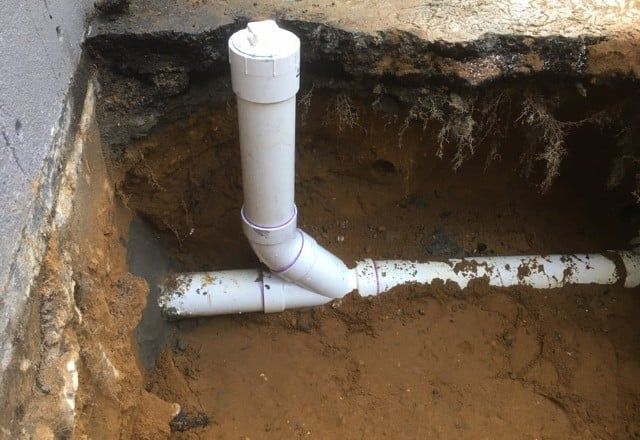 White PVC pipes in a dug-out trench, connected to a vertical pipe with a cap, near a concrete wall.
