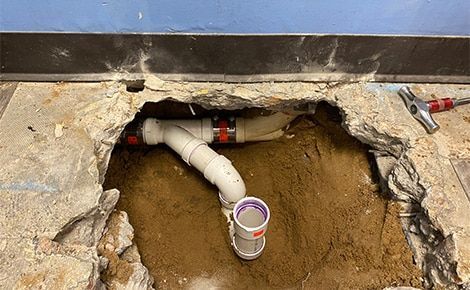 Hole in floor revealing plumbing pipes; white and red connectors, a partially visible cup, and a hammer.