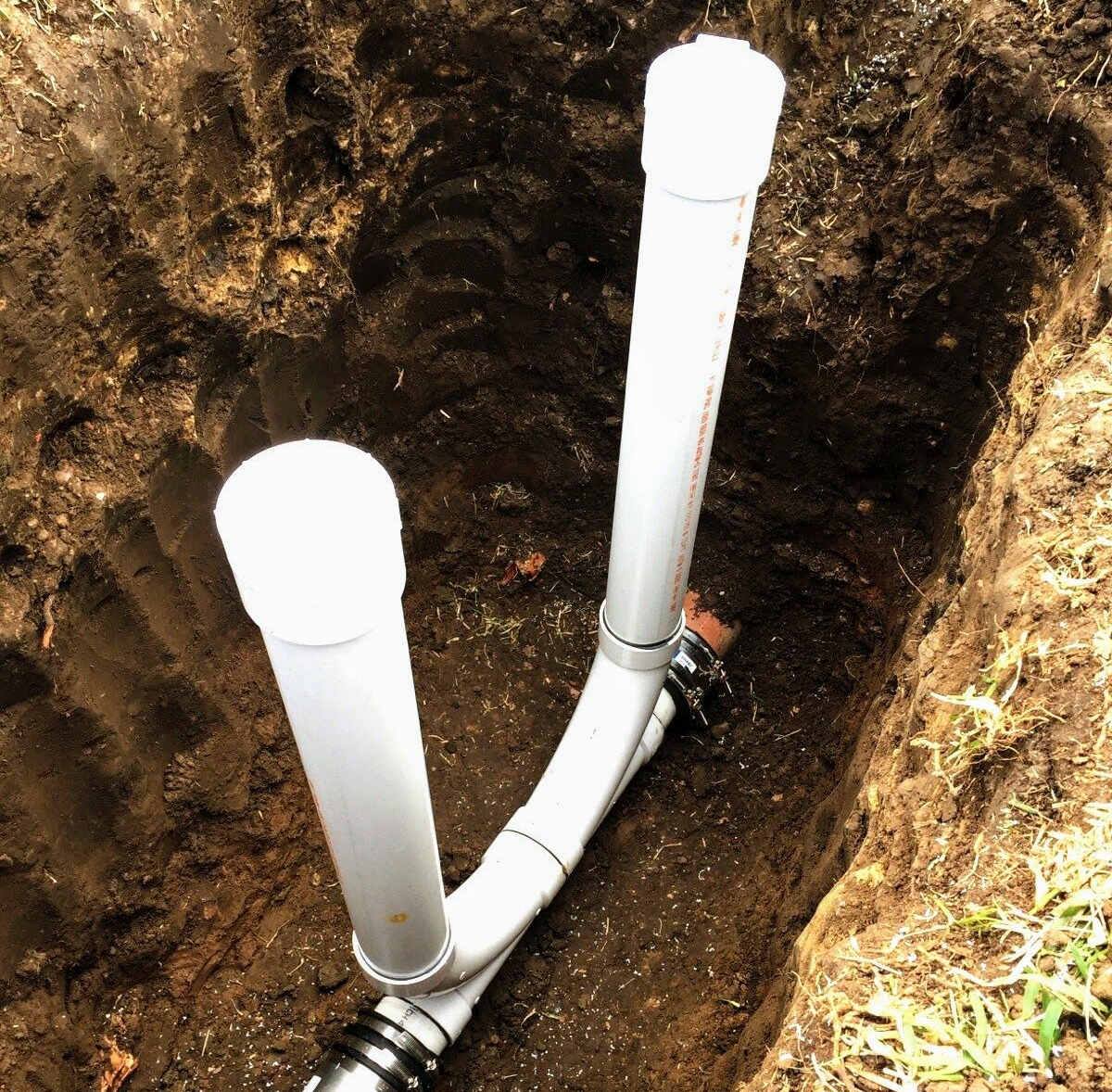 White PVC pipes in a soil trench, Y-shaped, capped. Outdoors.