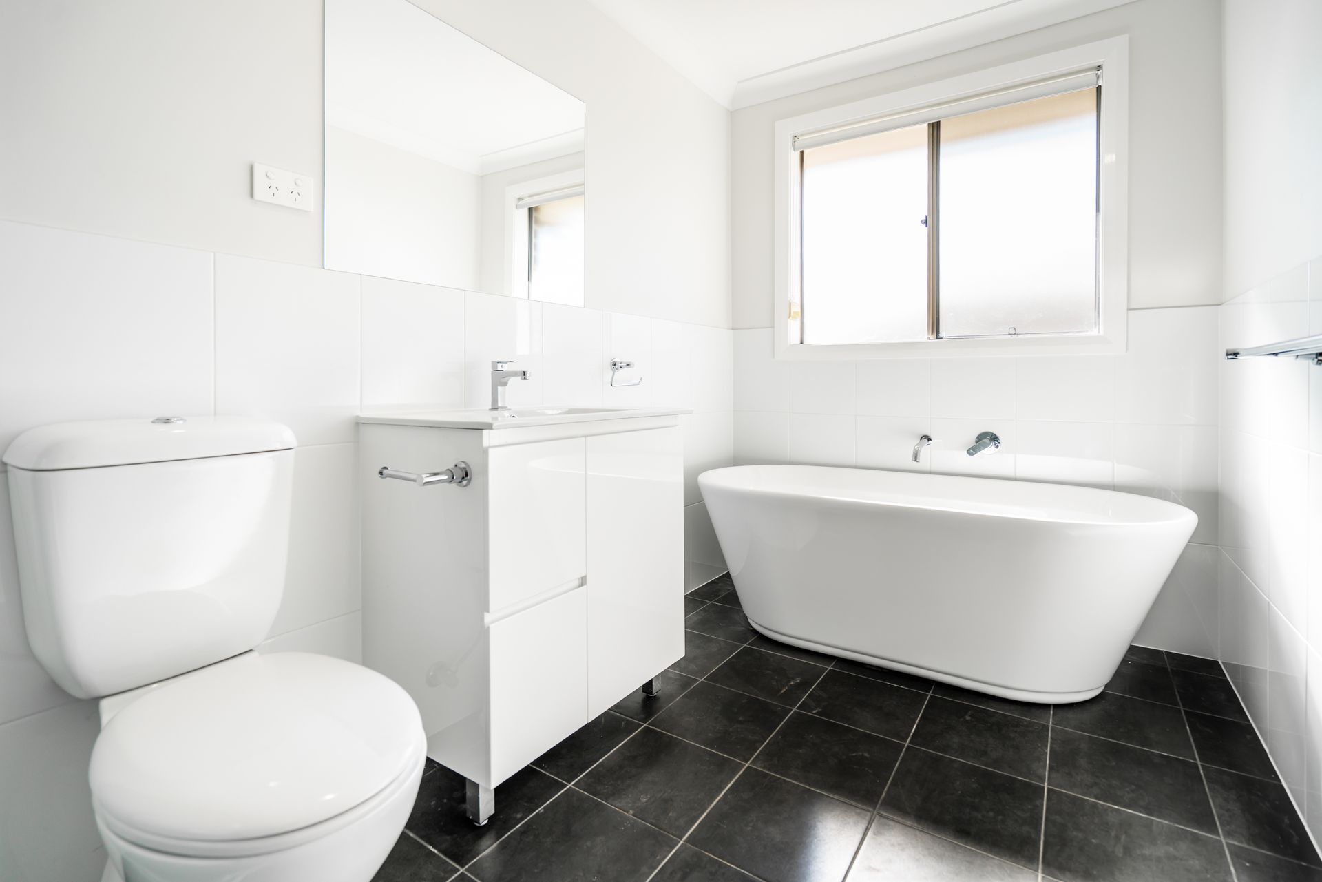 Modern white bathroom with black tile floor, toilet, vanity, and freestanding tub.