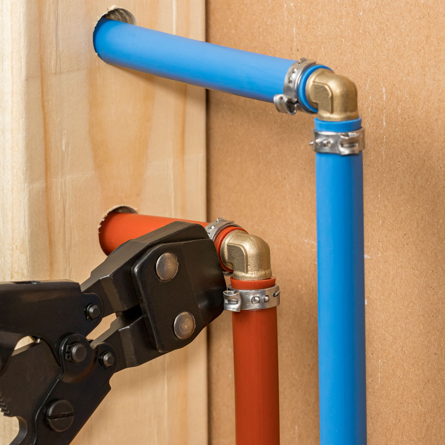 Plumber using crimp tool on PEX pipe with elbows, blue and red pipes, on a wooden frame.
