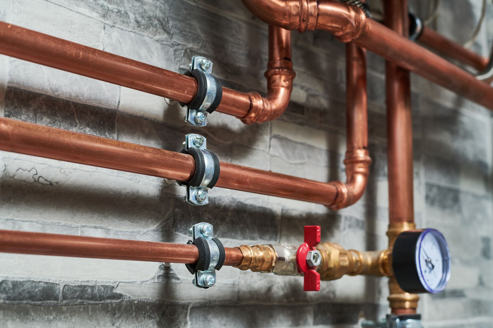 Copper pipes with black clamps on a textured gray wall, including a valve and pressure gauge.