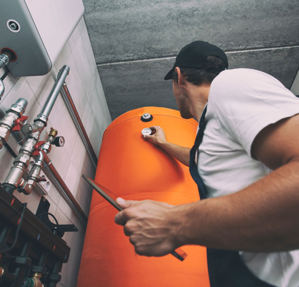 Man in work clothes checking a large orange water tank with a pointer in a utility room.
