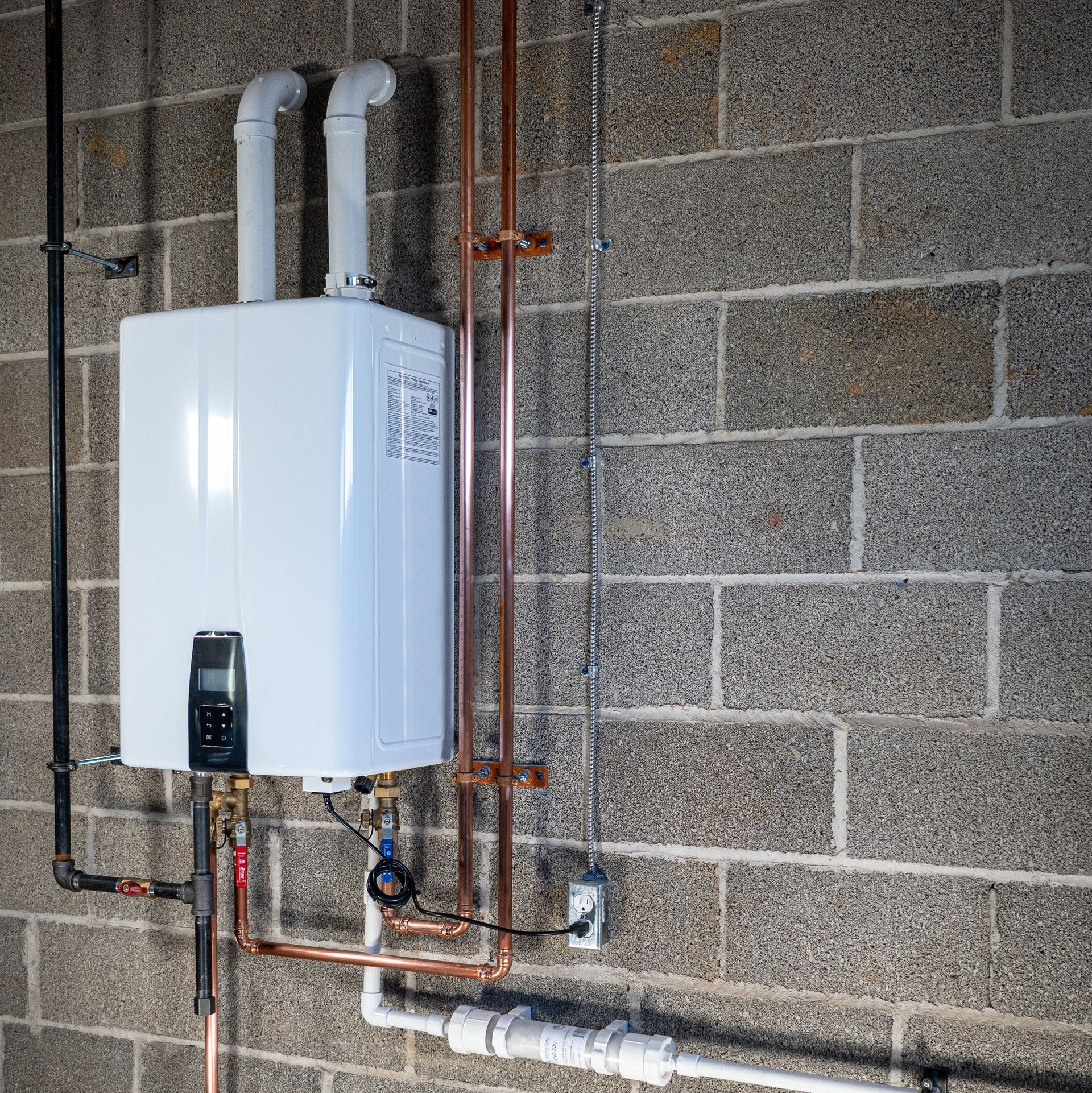 White tankless water heater on a gray brick wall with pipes and venting.