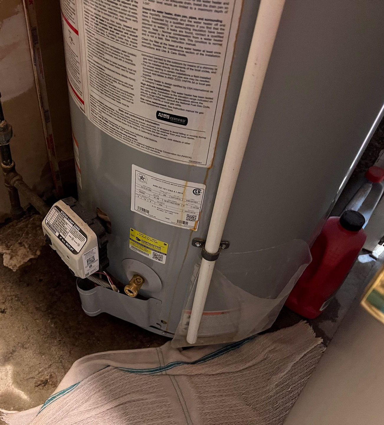 A water heater with a white pipe, standing on a concrete floor with a wet towel near it.