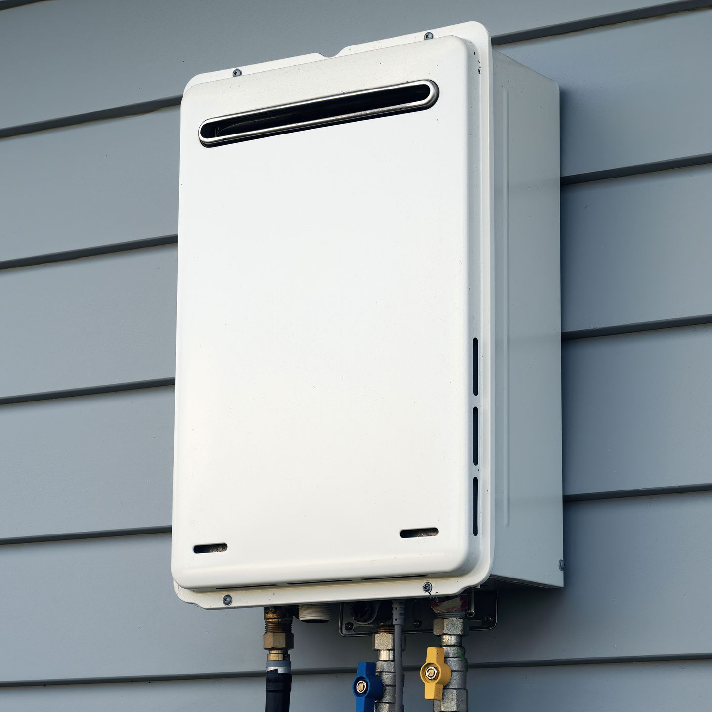 White tankless water heater mounted on gray siding, with water pipes below.