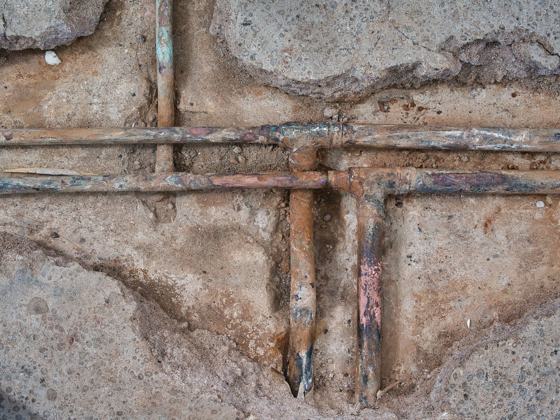 Corroded copper pipes embedded in cracked concrete. Brown and blue discoloration evident.
