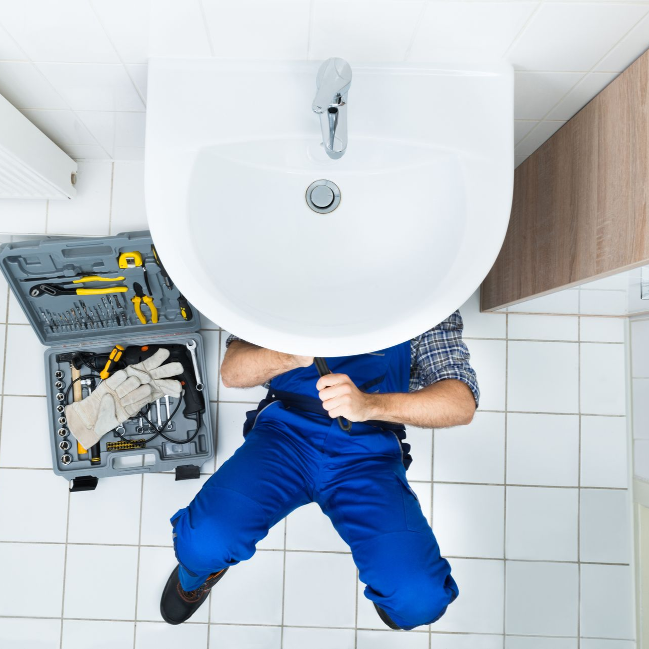 Plumber in blue coveralls working under a white sink with a toolbox on the tiled floor.