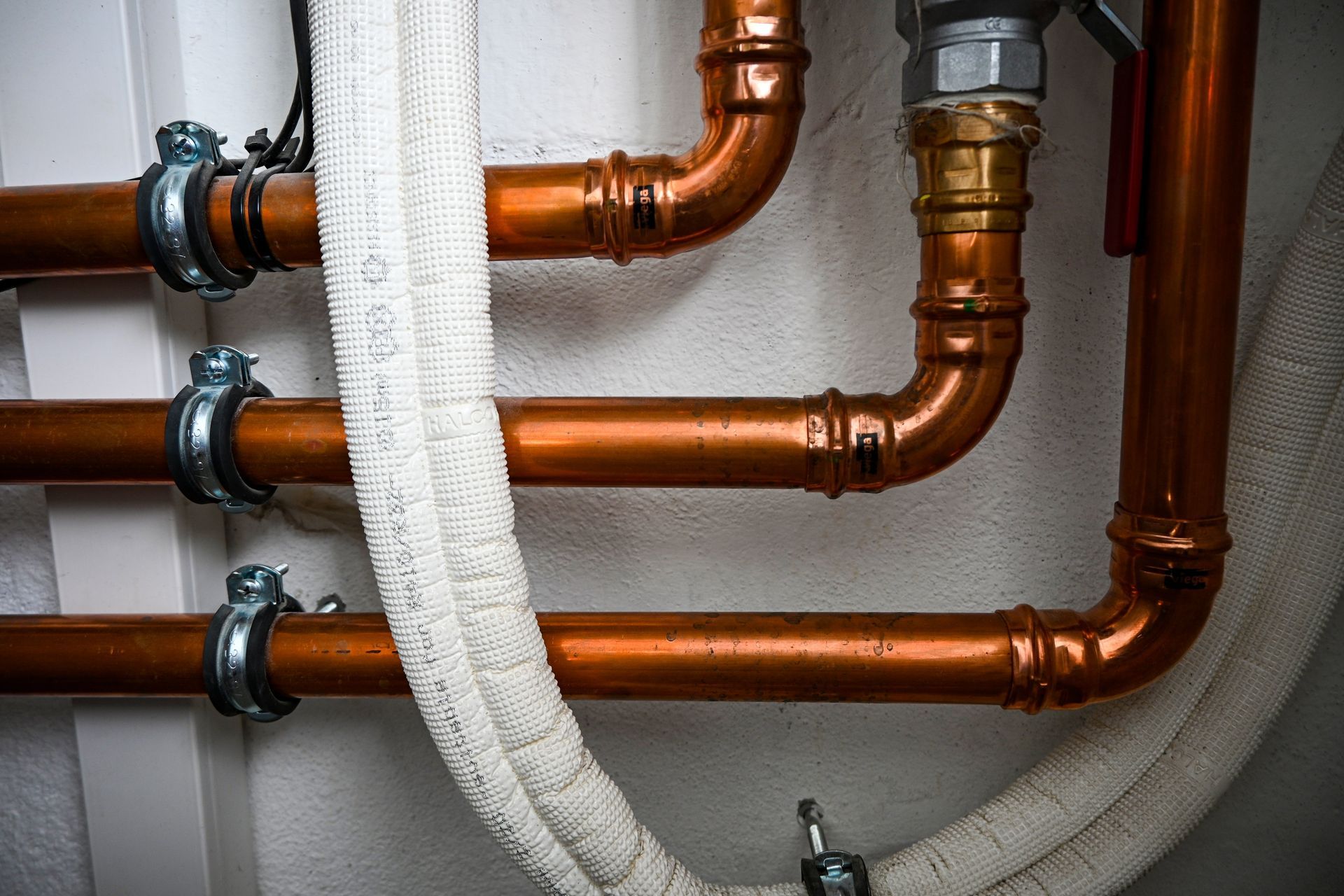 Copper pipes with fittings and insulation against a white wall.