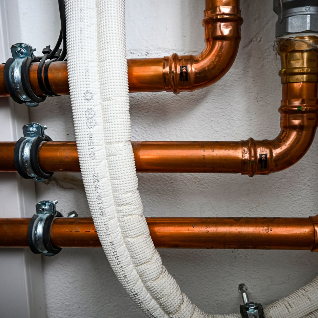 Copper pipes and insulated white tubing on a white wall, held by metal clamps.