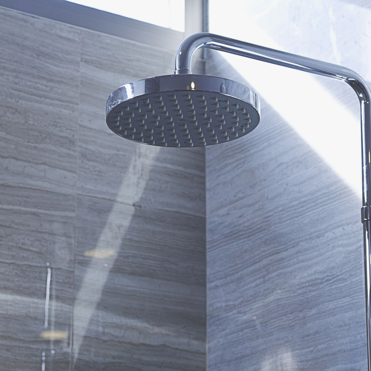 Chrome shower head on a silver bar against grey marble wall. Sunlight shines.