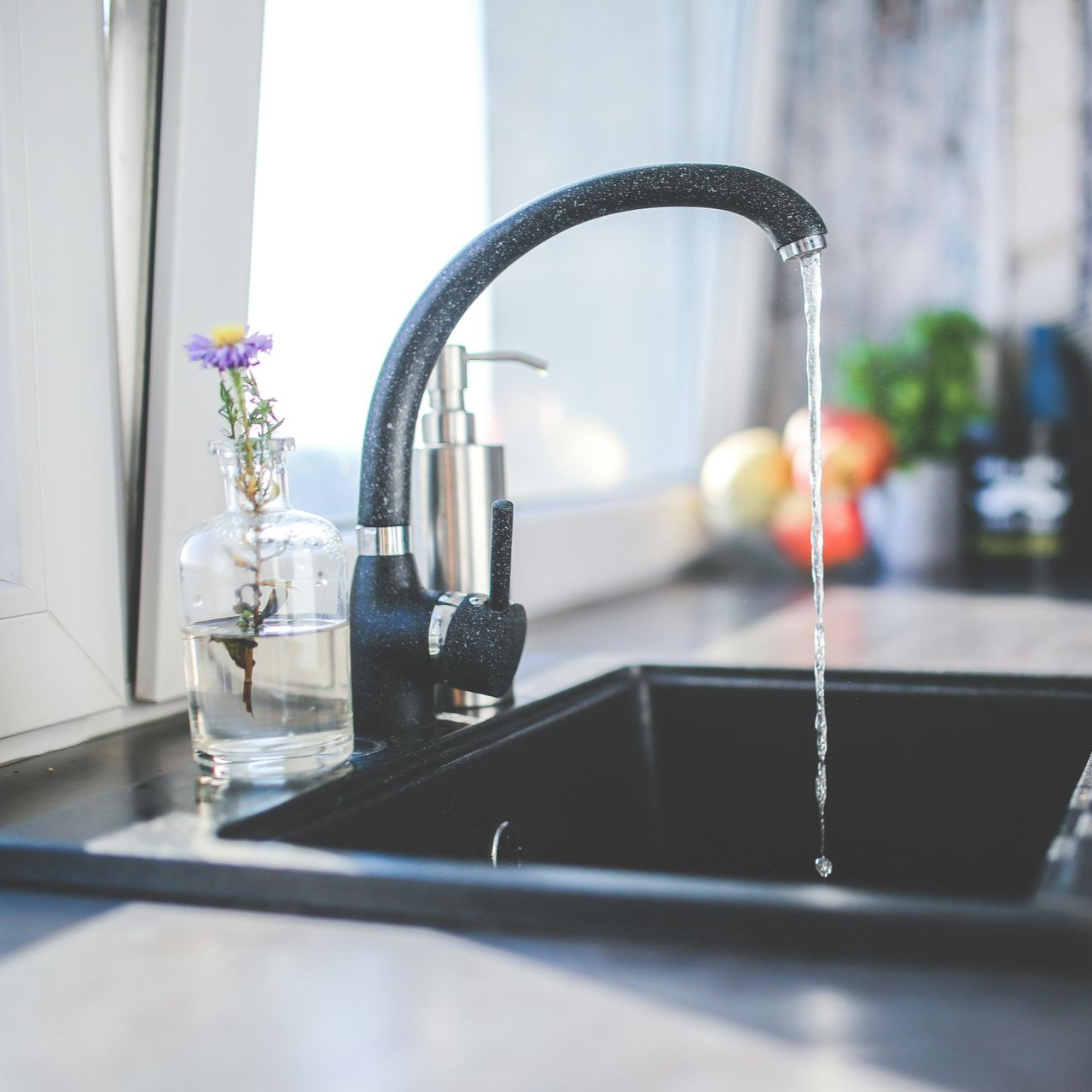 Water flowing from a black kitchen faucet into a dark sink. A vase with purple flower sits on the counter.