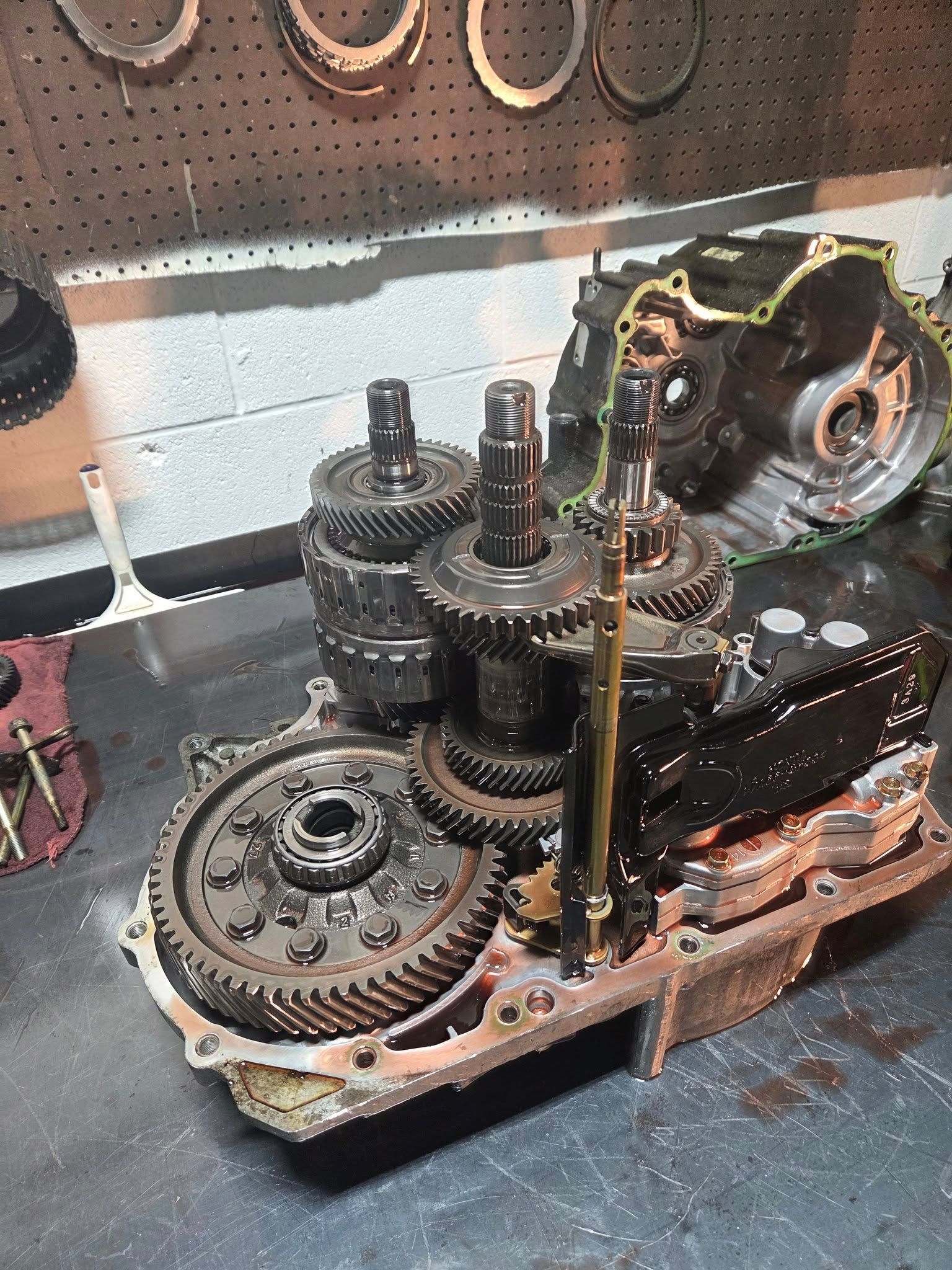 Disassembled car transmission parts on a workbench. Gears, shafts, and housing are visible in a garage setting.