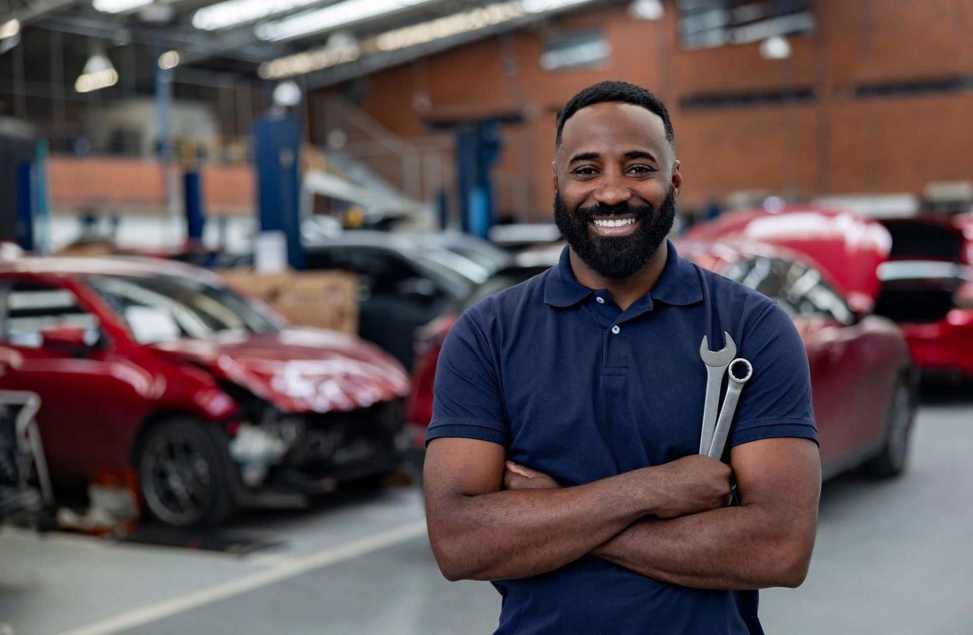 Mechanic in a blue shirt with a wrench in a car repair shop smiles, arms crossed, cars in background.