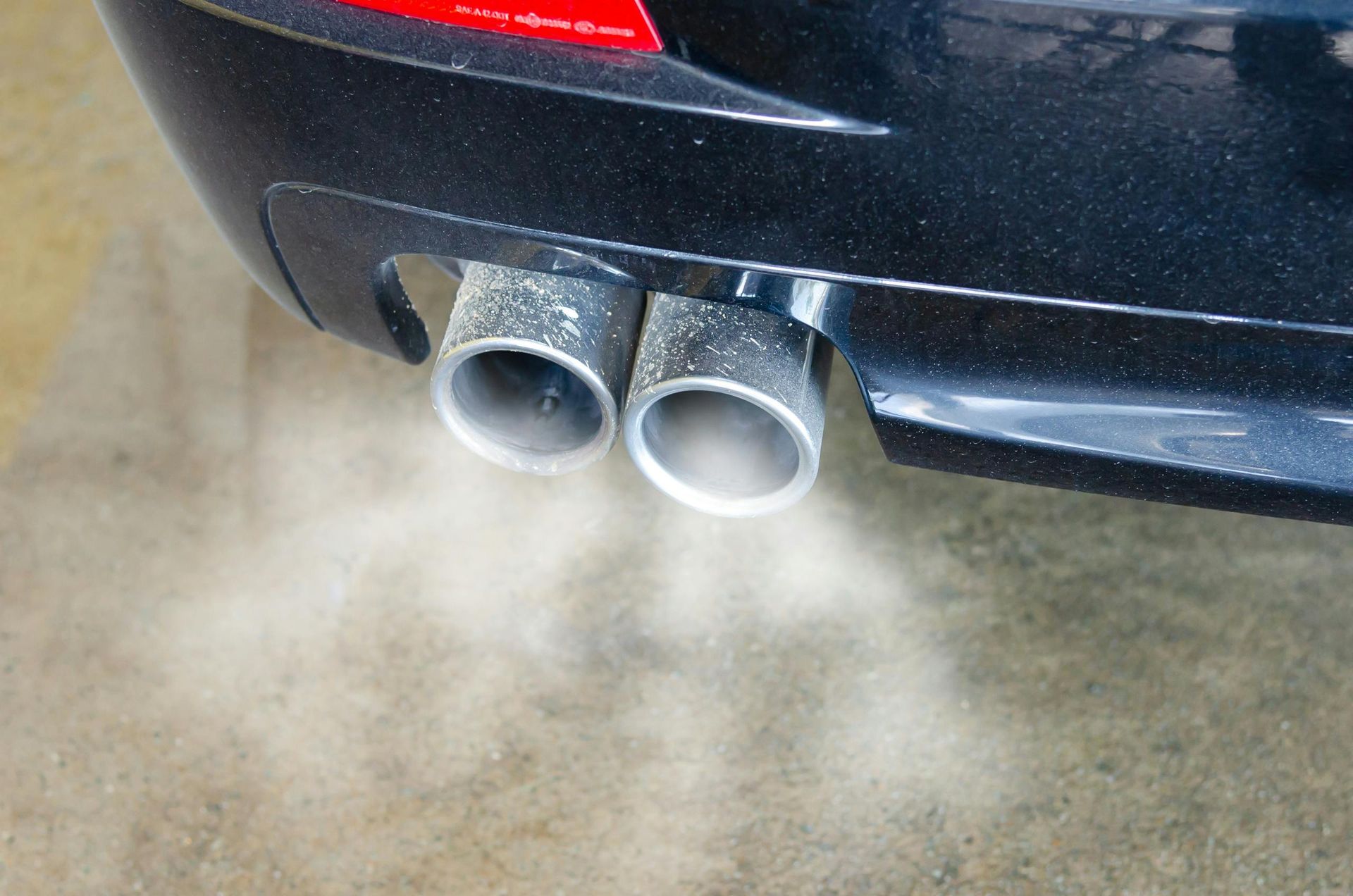 Close-up of a black car's exhaust pipes releasing white smoke onto a tan surface.