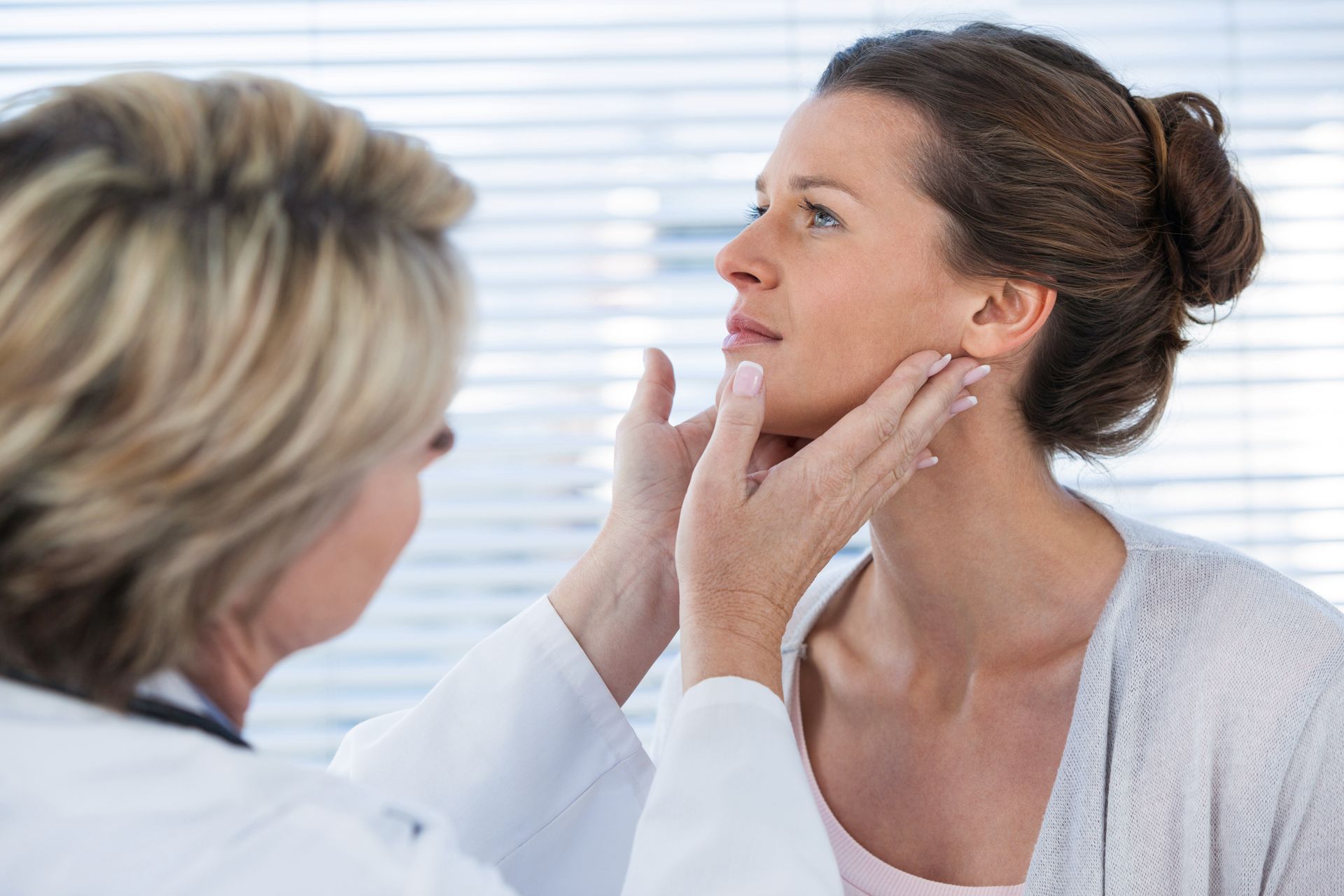 Doctor examining a woman's neck in an office. The doctor, in a white coat, touches the woman's jawline.