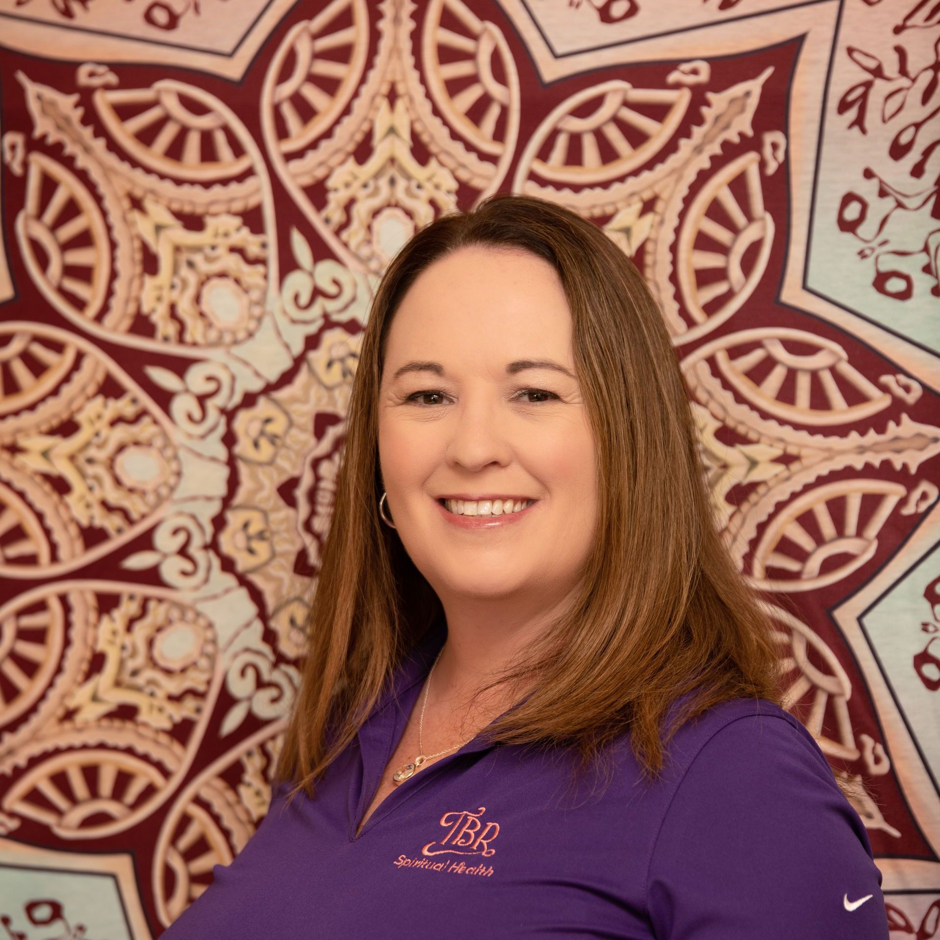 A woman in a purple shirt is smiling in front of a mandala tapestry.