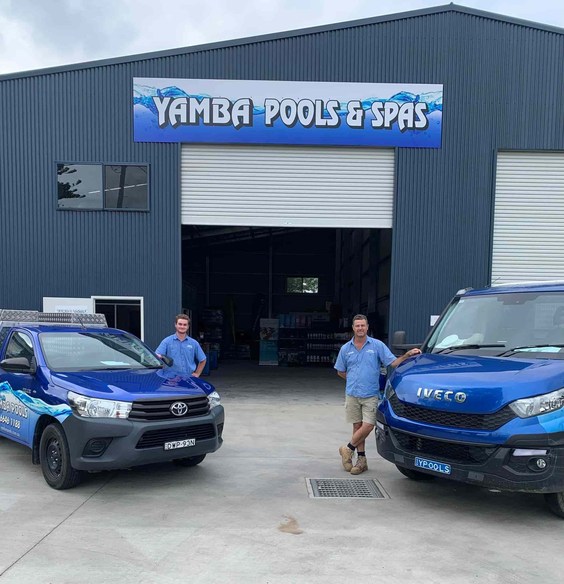 Two Men Are Standing In Front Of A Building With Two Blue Trucks Parked In Front Of It — Yamba Pools & Spas In Yamba, NSW