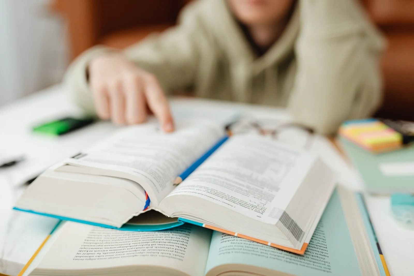A person in a light-colored hoodie sits at a white table, pointing to text in an open book resting on another larger book.