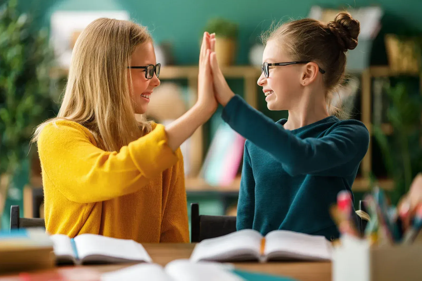 Two children with glasses high-five each other while sitting at a desk with open books in a classroom setting.