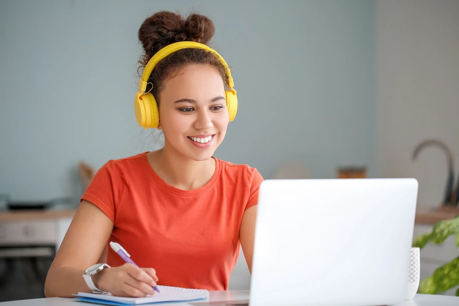 A person with hair in two buns wears yellow headphones, smiling while taking notes in a kitchen setting with a laptop.