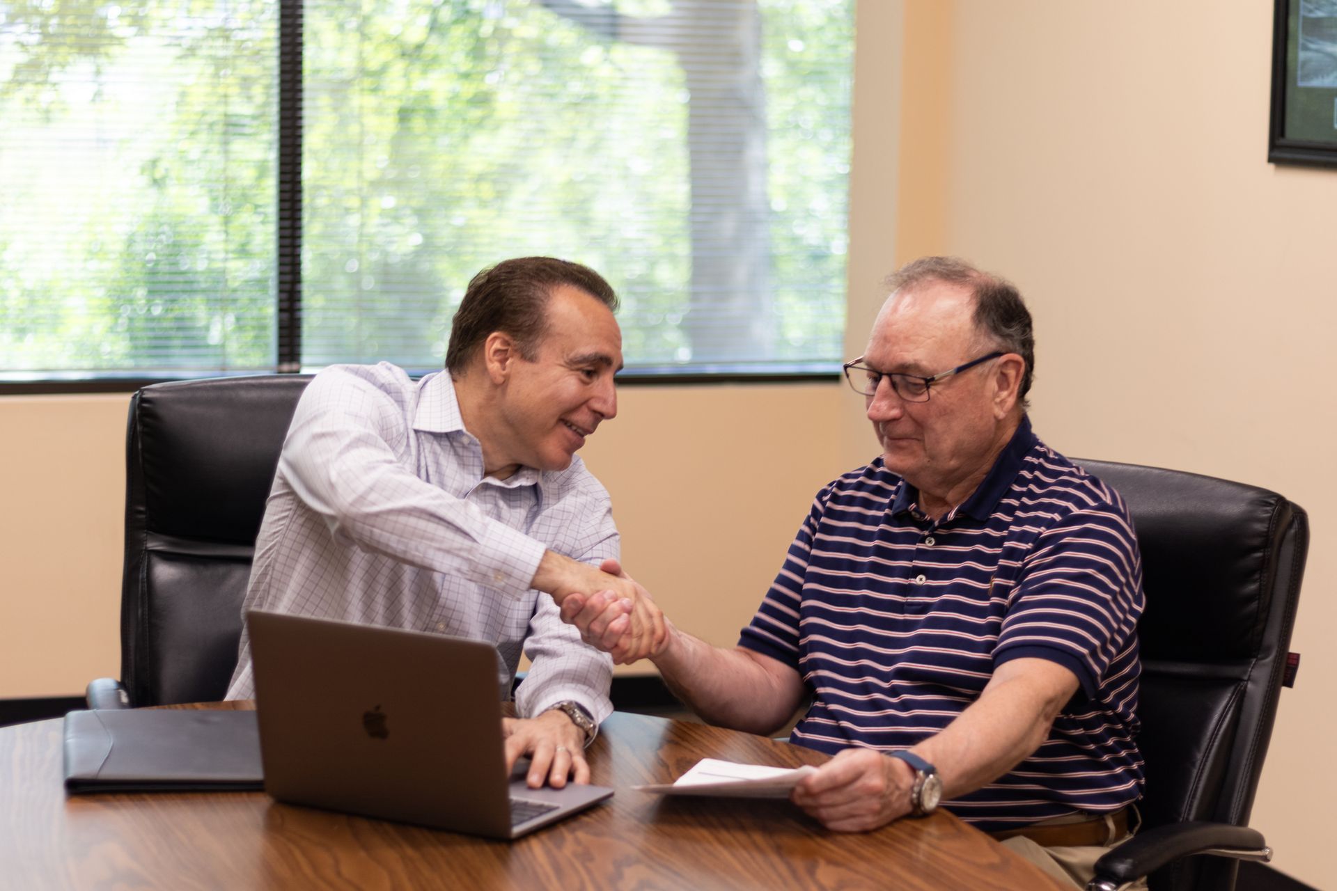 Two men are shaking hands while sitting at a table with a laptop.