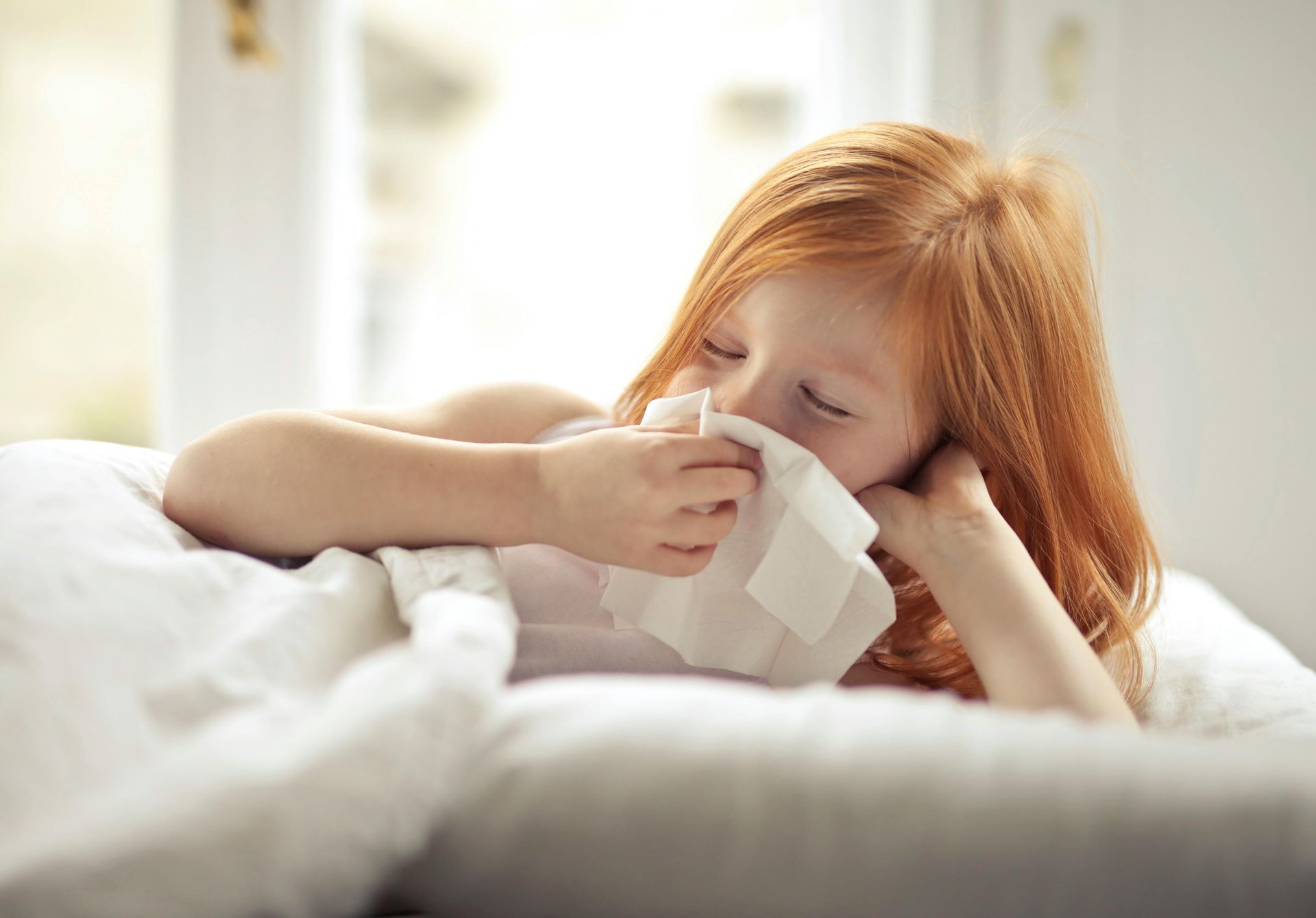 A little girl is blowing her nose with a napkin while laying on a bed.
