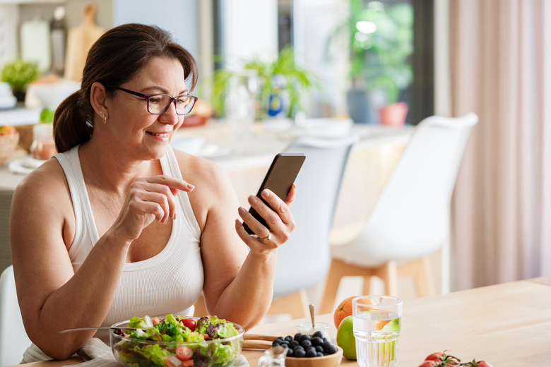Woman smiling at a kitchen table, tapping a smartphone beside a salad and a drink.