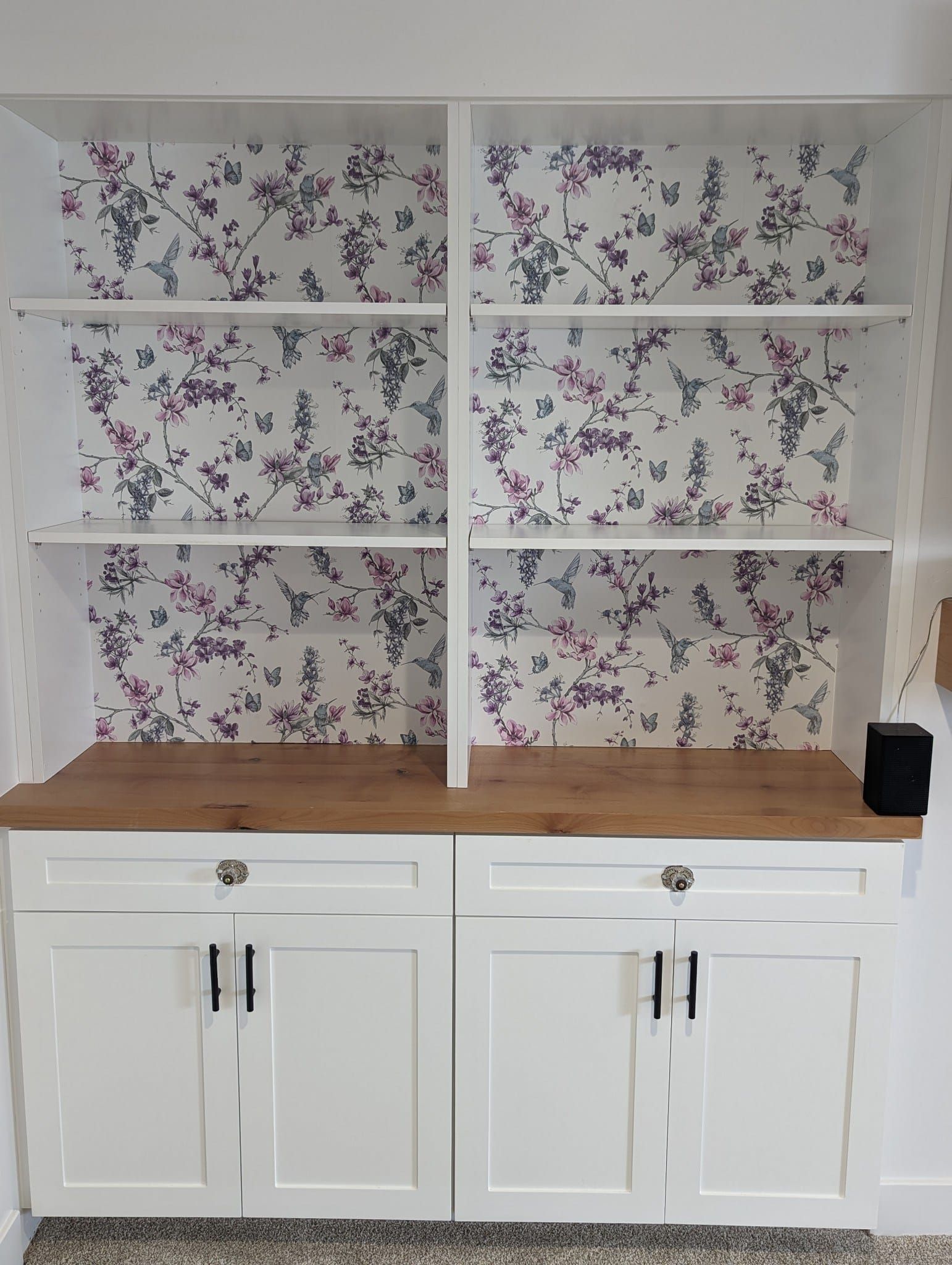 A bookshelf with floral wallpaper on the back of it.