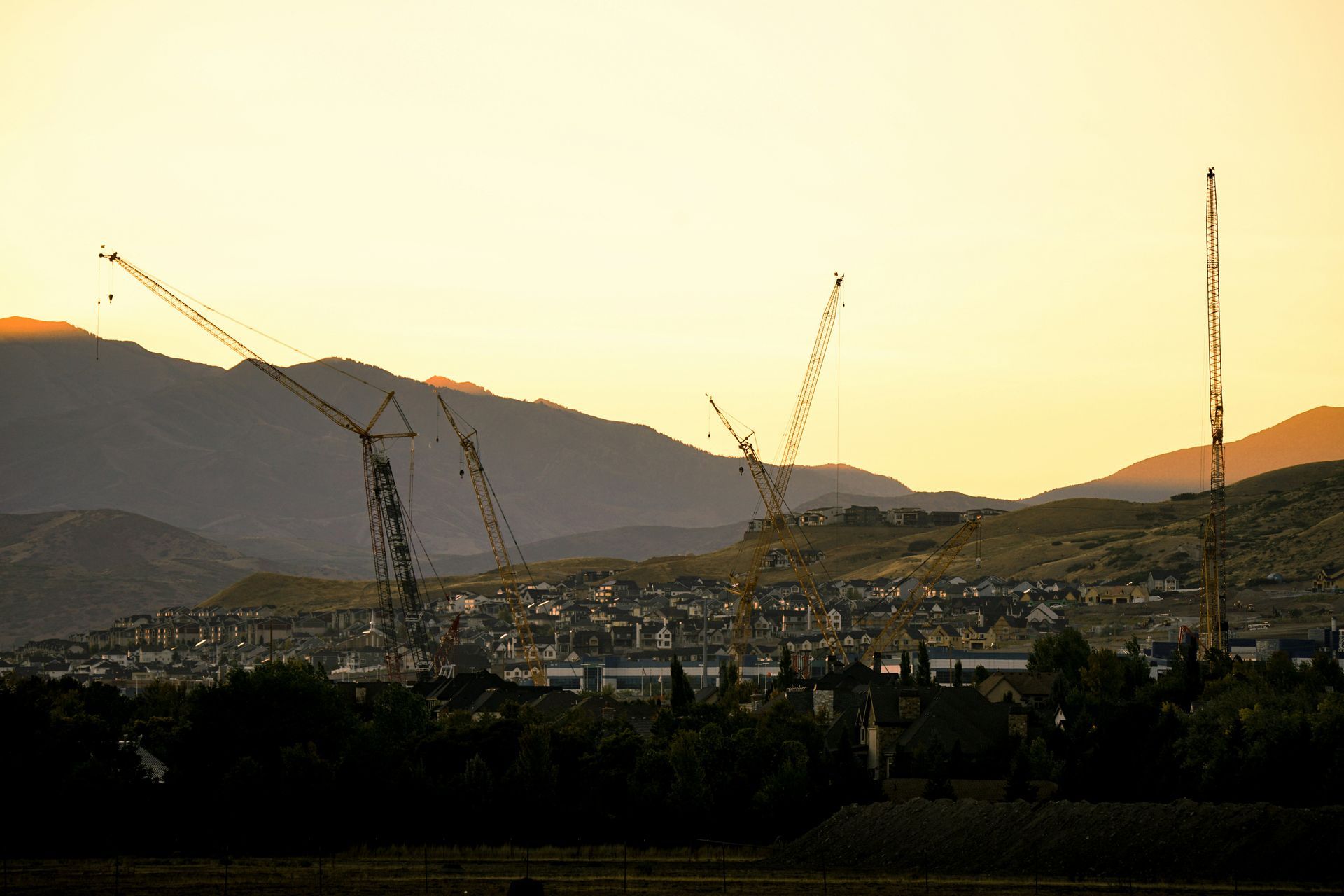 Cranes towering over a growing suburban neighborhood at sunset with mountains behind.