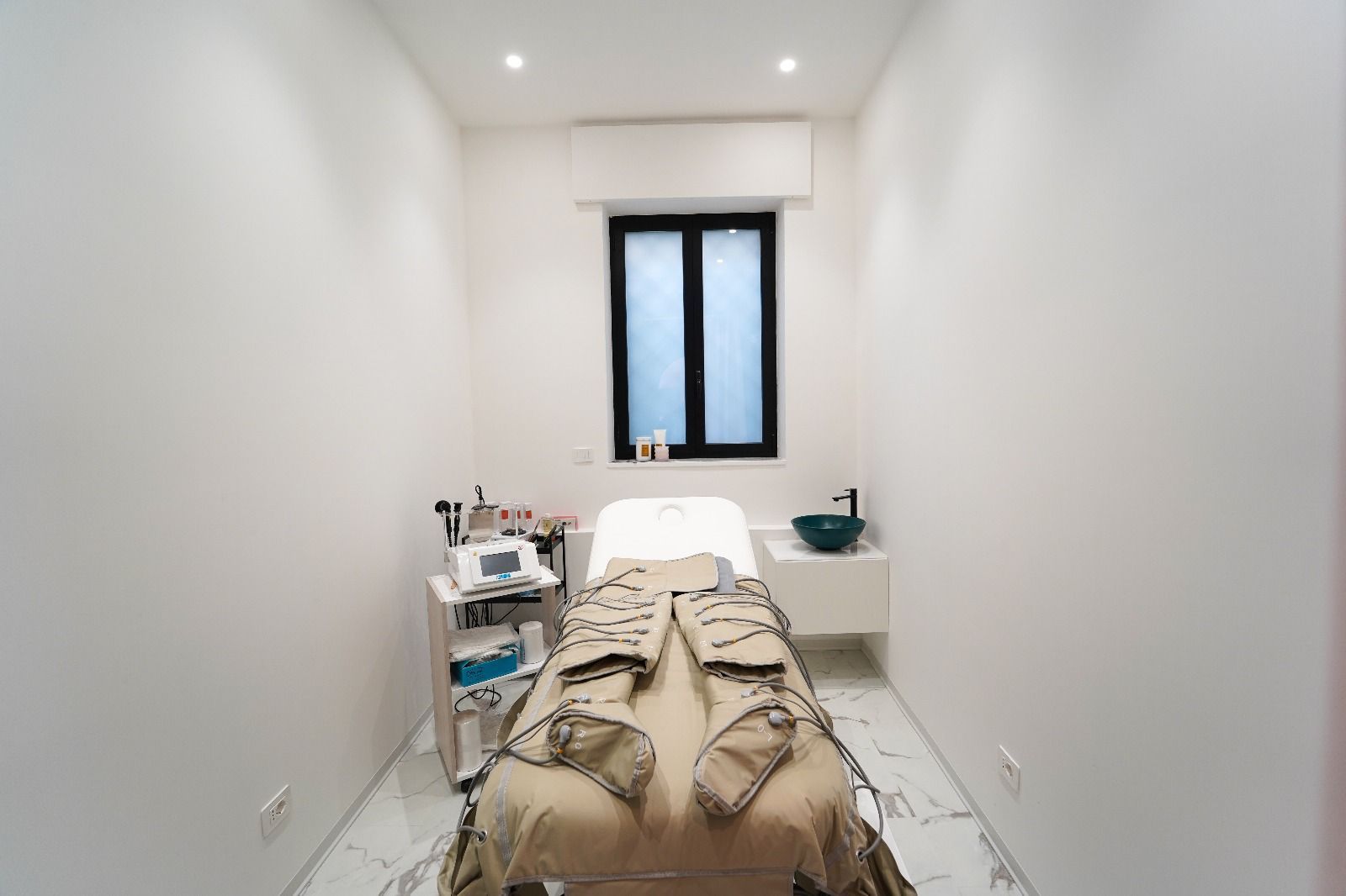 Piccola sala bianca per trattamenti con lettino e attrezzature. Sono visibili un lavandino e una finestra.