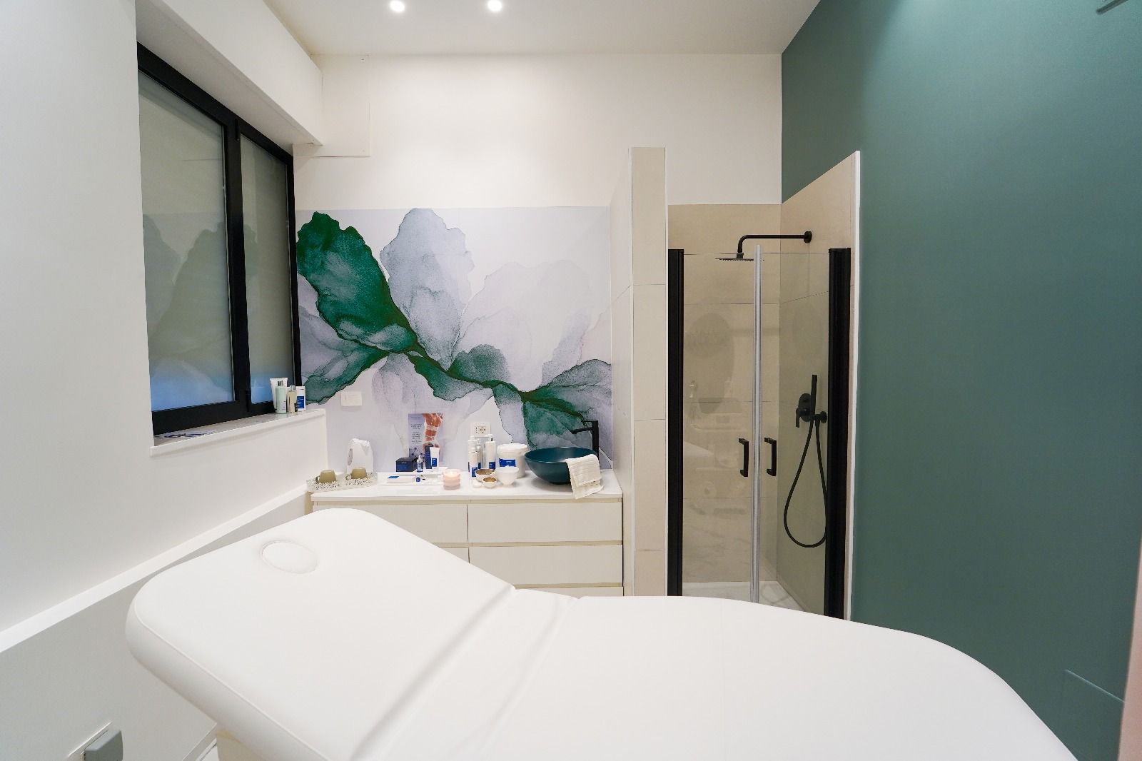 Sala per trattamenti spa con lettino per massaggi, doccia e parete dipinta con motivi astratti verdi.