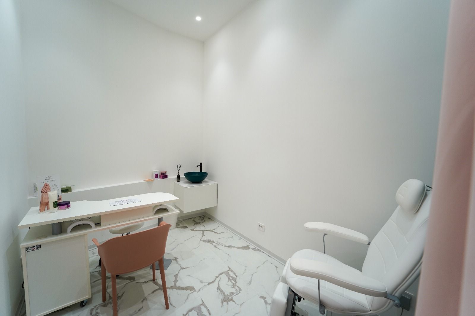 Una sala bianca per trattamenti di bellezza con scrivania, sedia, poltrona e lavandino.