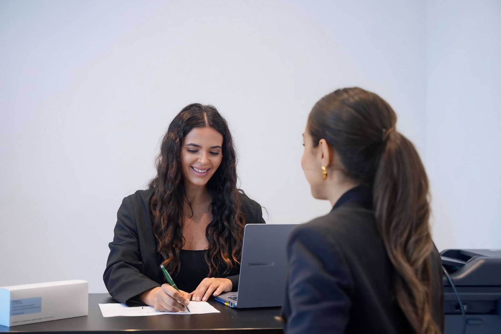 Donna che scrive alla scrivania, sorridente, di fronte a un'altra donna in un ambiente professionale con un computer portatile e dei documenti.