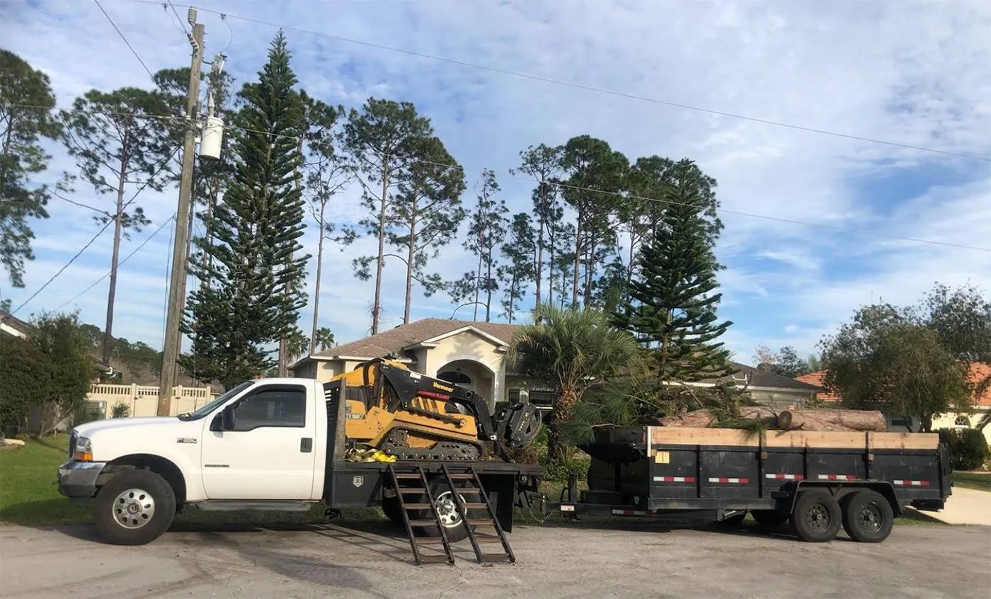 White truck with skid steer and trailer hauling tree debris parked in front of a house on a sunny day.