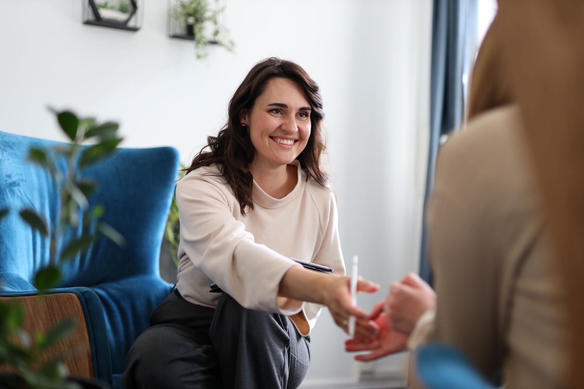 A smiling professional hands a stylus to a client in a bright, modern office with blue armchairs and indoor plants.