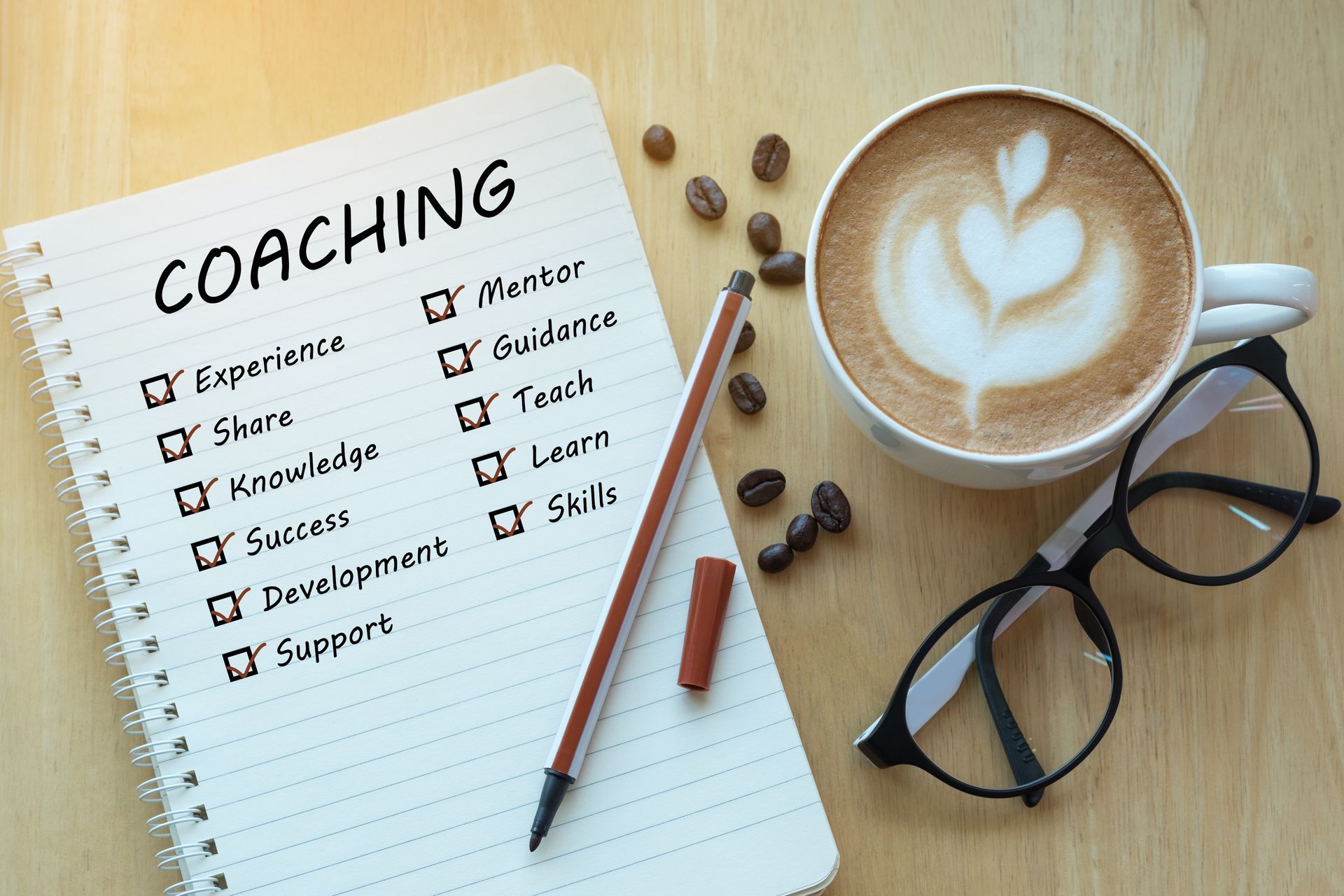 A notepad with a coaching checklist, a pen, coffee, and glasses on a wooden table.