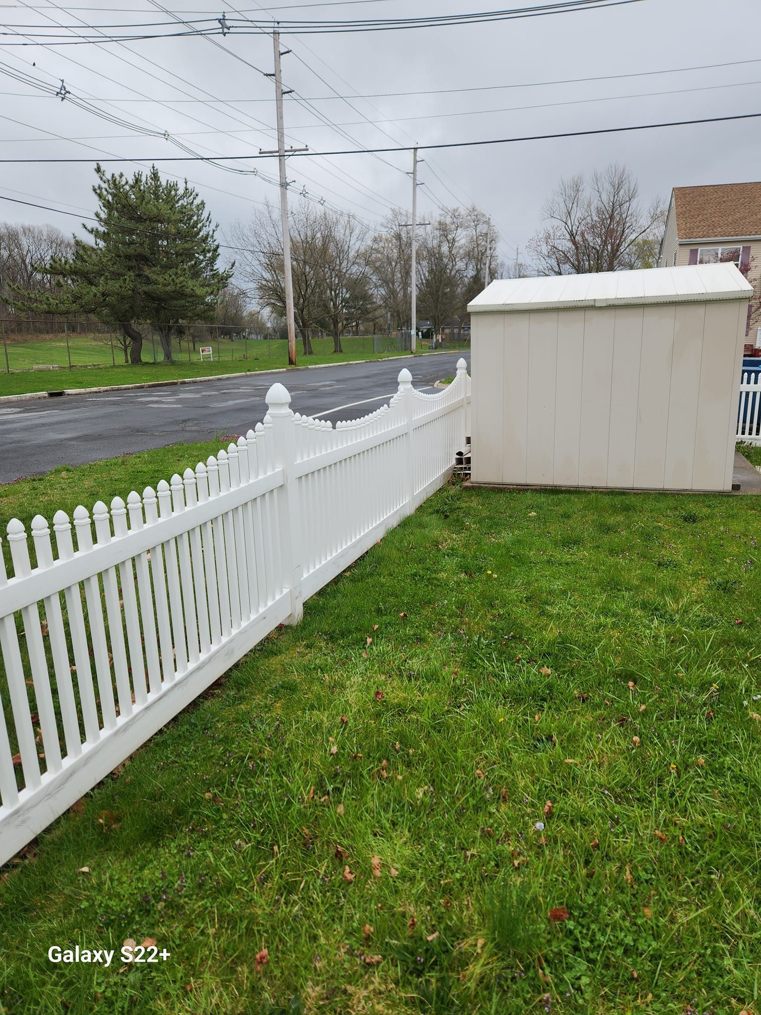 A white picket fence surrounds a grassy yard next to a road.